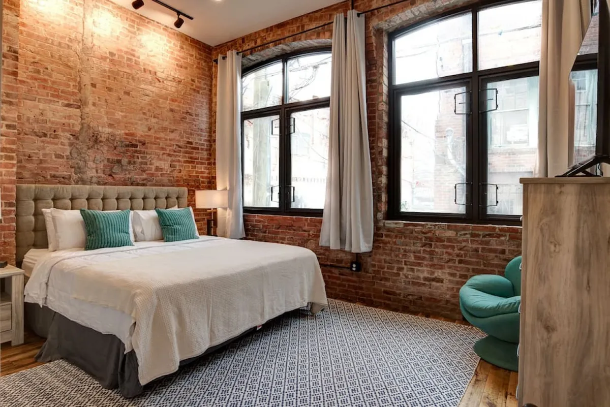This image features a cozy bedroom with a striking exposed brick wall, large windows letting in ample natural light, a stylish bed adorned with decorative pillows, and a green accent chair positioned for reading or relaxation. The combination of historic architectural elements and modern decor evokes a sense of both heritage and contemporary comfort.