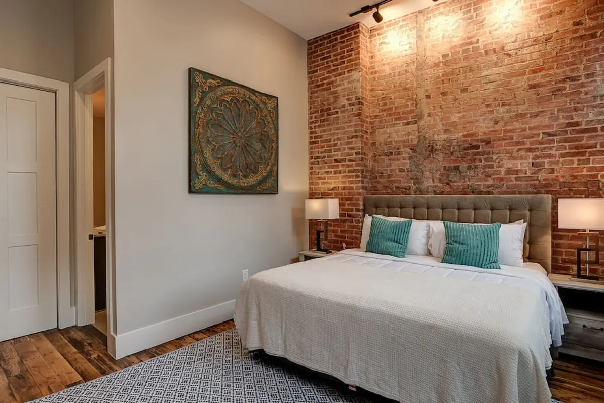 This photo shows a tranquil bedroom with a rich brick wall, a neatly made bed, decorative artwork, and matching nightstands. Natural light filters in, highlighting both the historic character and the contemporary styling of the space, suggesting an inviting retreat in the heart of the city.
