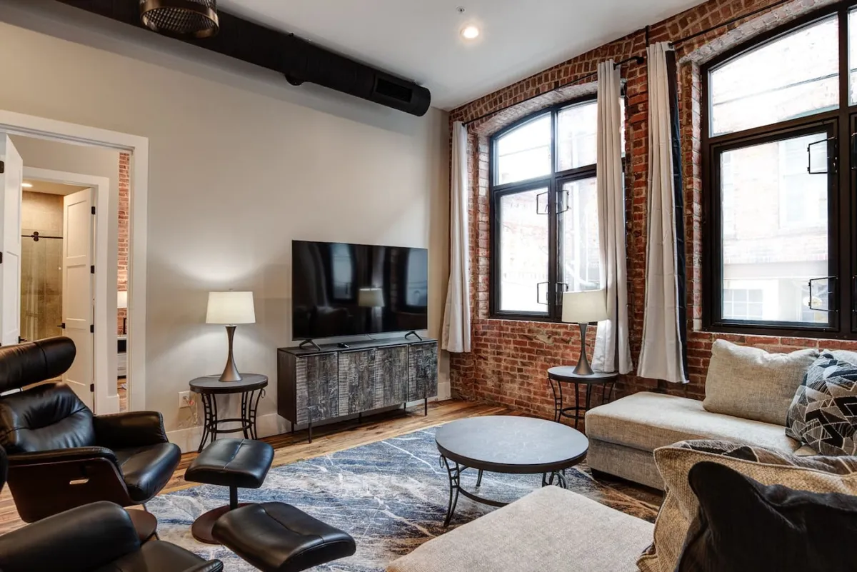 A welcoming living area defined by a striking exposed brick wall runs alongside large windows that flood the room with natural light. A cozy sofa and modern coffee table invite relaxation, while the TV and soft furnishings set the stage for socializing or unwinding after a day in the city.