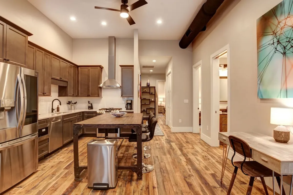 An open kitchen with a stylish island and bar stools, complemented by stainless steel appliances and warm wooden flooring. Colorful wall art adds a lively, personal touch. The layout encourages both cooking and casual entertaining, perfect for hosting friends or enjoying spontaneous meals together.