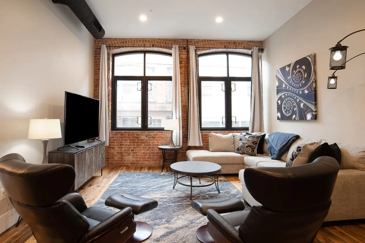 This image showcases a bright, open living area with large windows that allow natural light to pour in, a comfortable sofa perfect for group gatherings, modern artwork adding to the stylish ambiance, and a TV unit for entertainment. The view outside hints at the downtown location, emphasizing the connection between the condo's interior and the vibrant city just steps away.